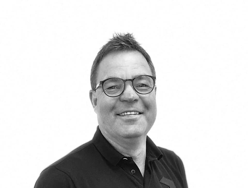 Sort/hvid portræt af en smilende midaldrende mand iført briller og sort skjorte mod en hvid baggrund.