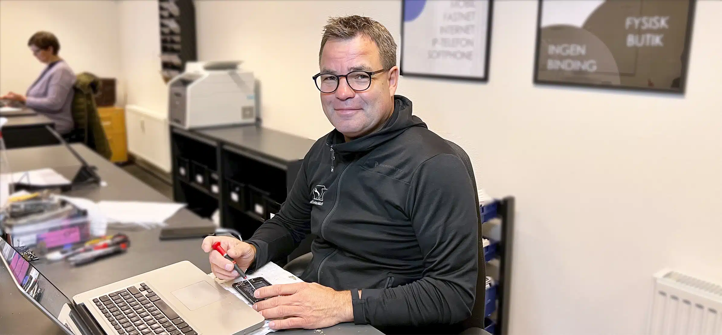 Mand i briller smiler til kameraet, mens han sidder ved et skrivebord og udfører iPhone reparation Viborg med en bærbar computer og papirer, med en anden person, der arbejder i baggrunden.