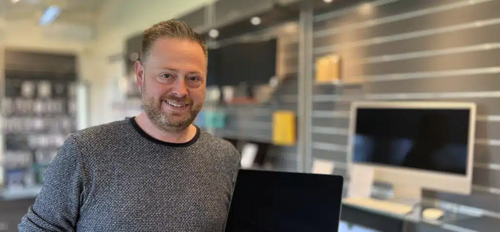 En smilende mand med skæg stående i et moderne kontor eller butik med computere og hylder i baggrunden.