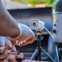 En person skærer kogt kød foran et webkamera på et stativ og en mikrofon med en grill synlig i baggrunden.