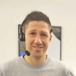 En smilende mand med kort hår iført et headset i kontormiljø.